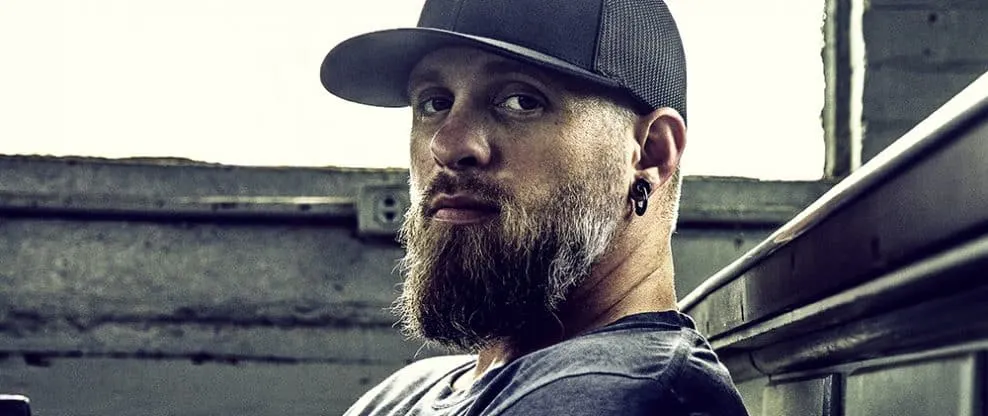 Bearded man wearing black cap and earrings, leaning on a windowsill in an urban setting.