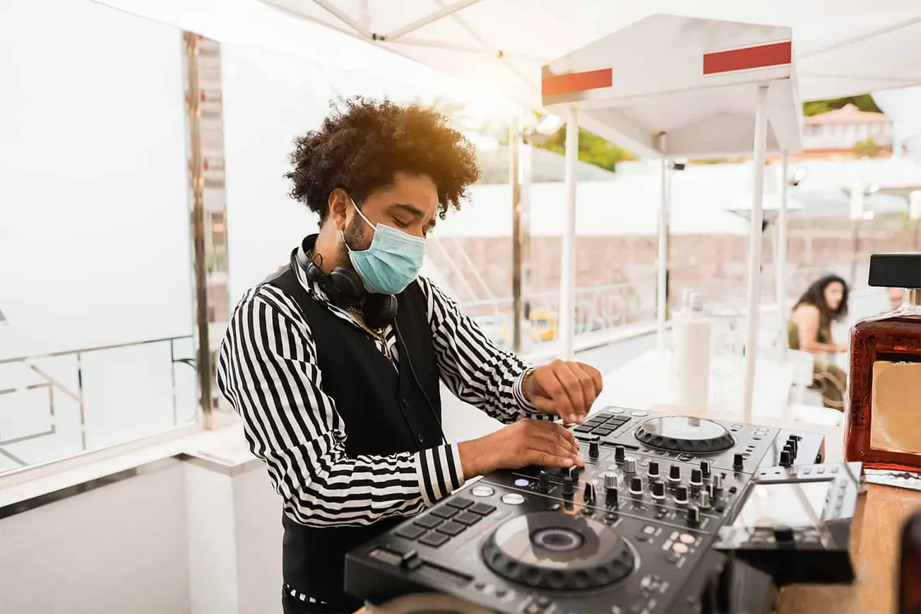 Young DJ wearing face mask operating turntables at outdoor music event, promoting safe live music experiences, blending DJ cu
