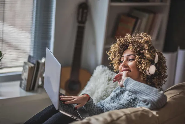 Young woman with curly hair wearing headphones and using a laptop in a cozy home setting, enjoying music or a podcast, with a