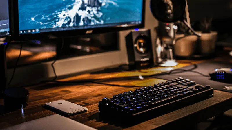 A modern desktop setup featuring a computer monitor, mechanical keyboard, smartphone, and audio equipment on a wooden desk in